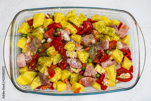 Raw vegetables ( potatoes, carrots, green peas, red pepper and onion) with pork meat garlic and spices in pyrex tray prepared for cooking in the oven.