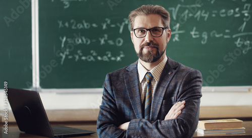 Portrait of smiling mature teacher in classic suit and eyewear standing cross-armed in classroom