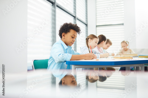 Pensive children wearing casual outfits sitting at school desk in modern classroom completing task in their notebooks, copy space