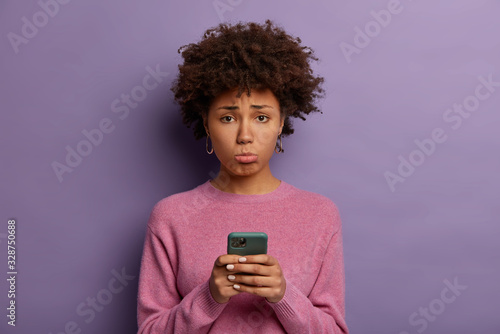 Unhappy disappointed woman with Afro hair, purses lower lip, holds smartphone, sad to miss chance of good shopping sale, upset not to receive call from boyfriend, poses indoor, dressed casually