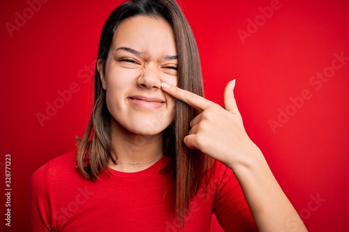 Young beautiful brunette girl wearing casual t-shirt over isolated red background Pointing with hand finger to face and nose, smiling cheerful. Beauty concept