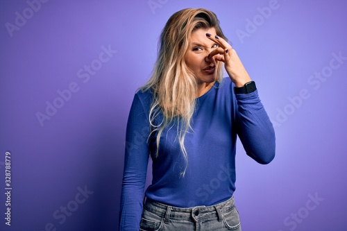 Young beautiful blonde woman wearing casual t-shirt over isolated purple background smelling something stinky and disgusting, intolerable smell, holding breath with fingers on nose. Bad smell