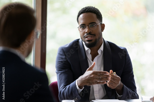 Confident concentrated African American male employee talk with colleague explain thought or idea, focused biracial businessman speak with coworker or partner, brainstorm at office boardroom meeting