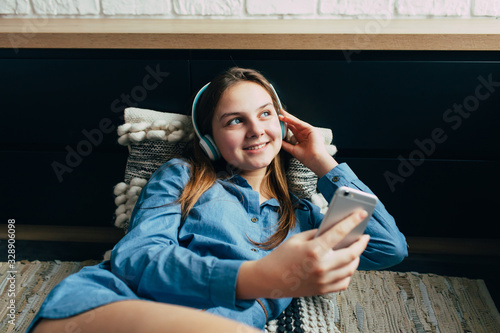 Smiling teenage girl with long hair lying at home interior and relaxing with cool music in headphone. Attractive student listening favourite songs via modern earphones using cellphone during morning