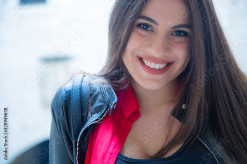 Bellissima donna italiana con un sorriso solare e capelli lunghi castani lisci indossa una camicia rossa e giacca nera di pelle in esterno di una città medievale