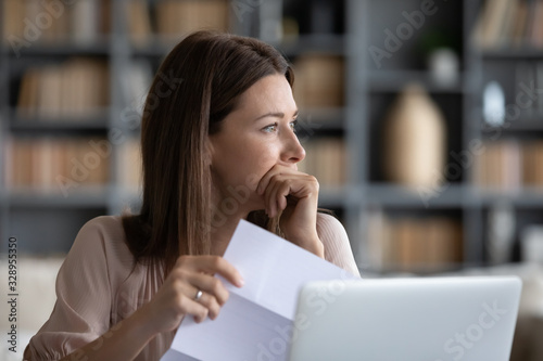 Head shot stressed young woman holding paper document, bank debt notification, thinking of financial troubles, looking away. Lost in negative thoughts depressed woman worrying about bad news notice.