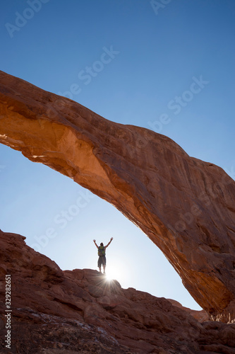 Backlit view of distant hiker raising his arms to the sky standing under a dramatic natural rock arch in front of golden sun