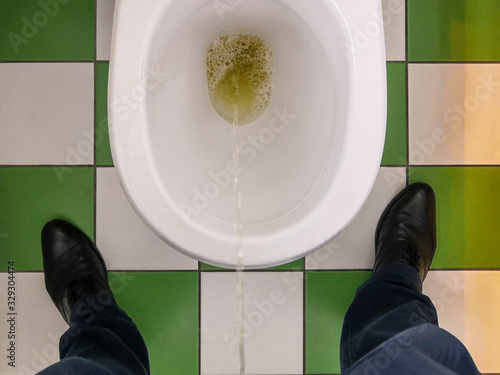 Top view of male feet and a toilet bowl into which a stream of urine flows. On the floor are white and green tiles