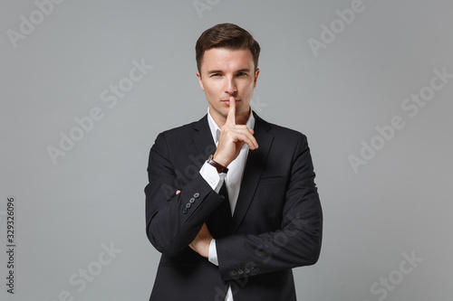 Secret young business man in classic suit shirt posing isolated on grey background. Achievement career wealth business concept. Mock up copy space. Say hush be quiet with finger on lips shhh gesture.