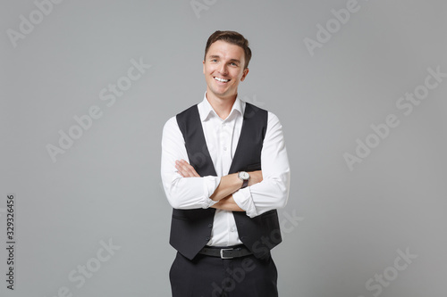 Smiling young business man in classic black waistcoat shirt posing isolated on grey wall background in studio. Achievement career wealth business concept. Mock up copy space. Holding hands crossed.