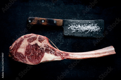 Raw Tomahawk steak dry aged with a meat cleaver on a dark stone background, top view