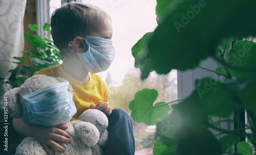 Quarantine, threat of coronavirus, virus protection, pandemic. Child and his teddy bear both in protective medical masks sits on windowsill inside house and looks out window. Focus on toy.