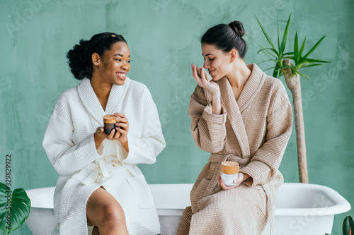 Cheerful pretty multiracial young ladies friends wear silk robes having fun hold glasses with champagne sit on bed in hotel, happy diverse girls bride and bridesmaid celebrate bachelorette spa party