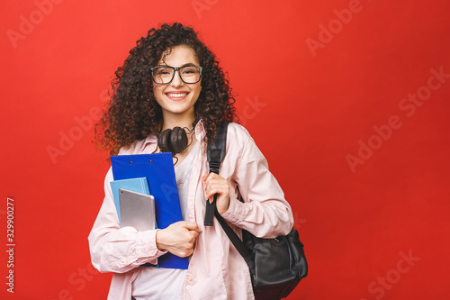 Young curly student woman wearing backpack glasses holding books and tablet over isolated red background.