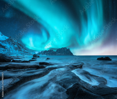 Northern lights above snowy mountains and sandy beach with stones. Aurora borealis in Lofoten islands, Norway. Starry sky with polar lights. Night winter landscape with aurora, sea with blurred water