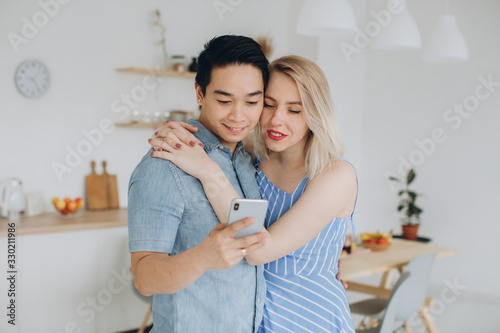 Asian man and his caucasian blonde woman spend time together in kitchen and looking at smartphone
