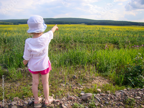 Child shows with index finger to the yellow field, the forest. View from the back. Sky with clouds. Warm weather in summer.