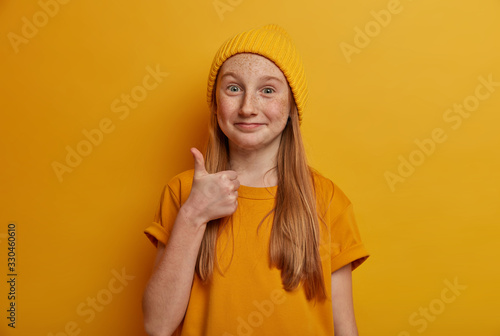 Portrait of freckled girl shows thumb up, agrees with something and says okay, like gesture, looks positively, wears yellow clothes in one tone with background. Children, body language concept