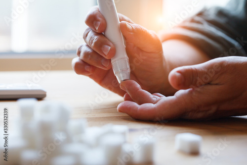 senior woman hands using lancet on finger at home to check blood sugar level, glucometer and sugar cubes on wooden table close up, diabetes concept, elderly health care, sunny morning