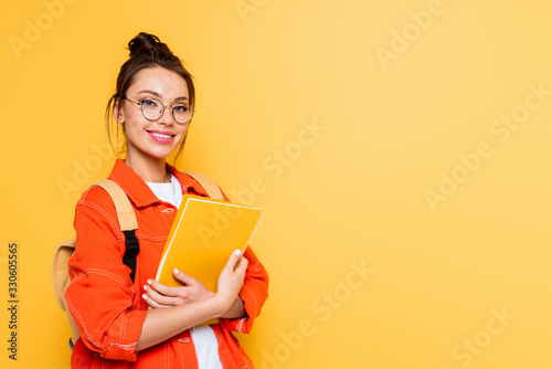 cheerful student in glasses looking at camera while holding notebook isolated on yellow
