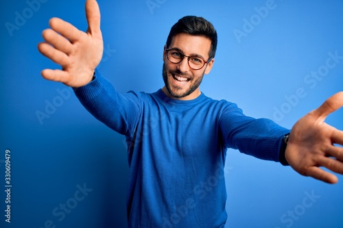 Young handsome man with beard wearing casual sweater and glasses over blue background looking at the camera smiling with open arms for hug. Cheerful expression embracing happiness.