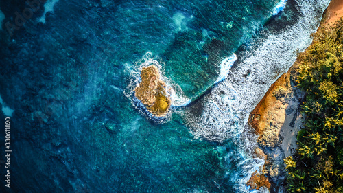 Aerial drone photo of a tiny island in a turquoise transparent water of an ocean, surrounded by sandy beach and palm trees of a tropical resort. View from the top