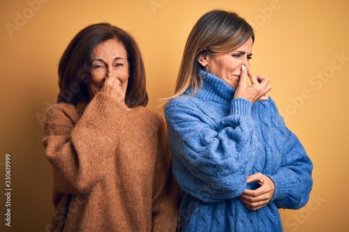Middle age beautiful couple of sisters wearing casual sweater over isolated yellow background smelling something stinky and disgusting, intolerable smell, holding breath with fingers on nose.