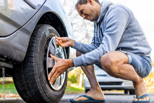 Missing cap cover on parked car tire wheel with man replacing hubcap with new shiny metal outside with difficulty