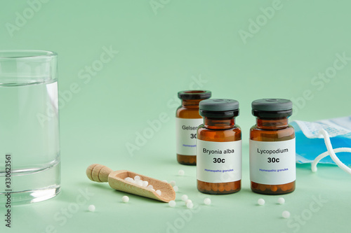 Bottles of homeopathic drugs - Bryonia, Lycopodium, Gelsemium - recommended by homeopaths for treatment and prophylaxis of Coronavirus Covid-19, glass of water, scoop, medical protective mask.