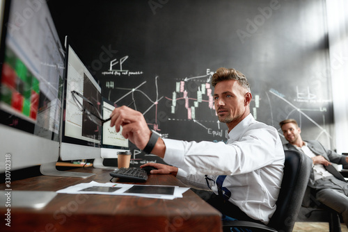 Be educated. Middle-aged trader sitting by desk in front of multiple computer monitors while working in the office. His colleague and blackboard full of data analyses in background.