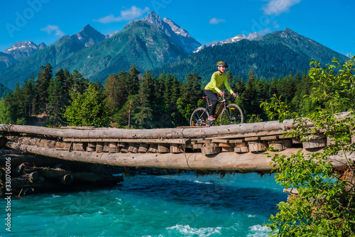 A young smiling girl on a cyclocross bike rides on a wooden bridge against a background of blue sky and high mountains