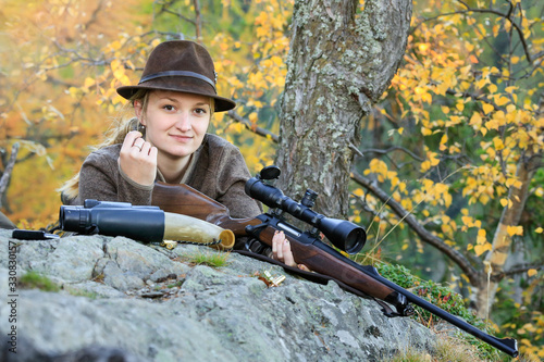 Young, Clever Woman Deer Hunter wearing green uniform with Rifle on the rock in Background autumn trees