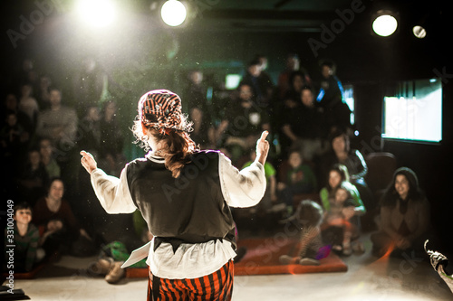 A performing arts entertainer is seen from the back in selective focus, dressed as a pirate on stage during a comedy act with blurry audience at back.