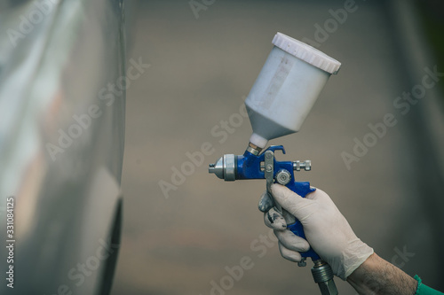 Caucasian man is spraying color with a compressed air paint gun on the vintage car as a restoration project. Man wearing protective equipment such as mask and gloves, professional automotive painter.