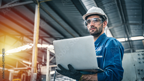 Confident engineer in blue jumpsuit holding laptop computer in a warehouse.