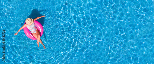 Zenith aerial view of a swimming pool in summer. Young girl in a swimsuit and hat floating with pink donut.