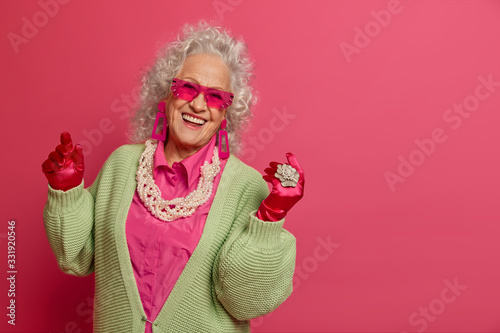 Joyful curly elderly woman dances carefree, has fun, wears trendy sunglasses, dressed in fashionable clothes, enjoys life, models against pink background, copy space for your advertisement or text