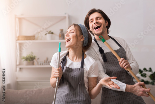 Cheerful Couple Using Mop Handles As Microphones, Having Fun During Spring-Cleaning