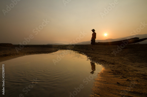 Young man standing near a drying river with a wooden boat at the place that once had been a big lake before, climate change, water crisis concept.