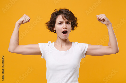 Strong young brunette woman girl in white t-shirt posing isolated on yellow orange background studio portrait. People sincere emotions lifestyle concept. Mock up copy space. Showing biceps, muscles.