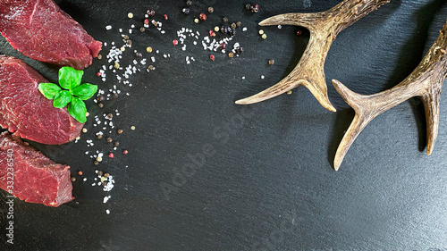 Roe deer steak with basil, pepper and salt on the dark background. In the corner is roe antlers as a decoration.