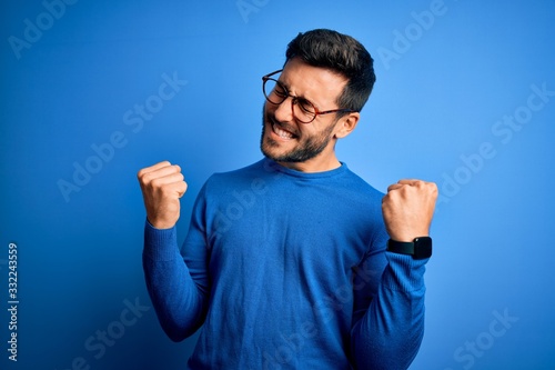 Young handsome man with beard wearing casual sweater and glasses over blue background very happy and excited doing winner gesture with arms raised, smiling and screaming for success. Celebration