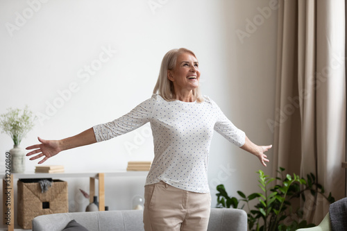 Overjoyed mature grandmother standing with outstretched arms near comfortable couch, breathing fresh air, enjoying freedom, happy life moment. Smiling older woman feeling thankful for good day.
