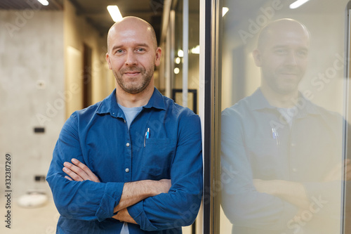 Waist up portrait of mature bald man smiling at camera while standing with arms crossed and posing confidently leaning against wall, copy space