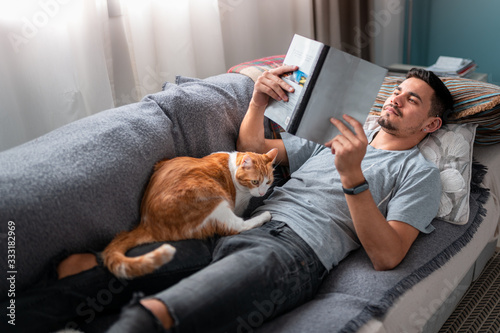 hombre joven acostado en un sofa lee un libro. Un gato blanco y marron descansa encima del hombre