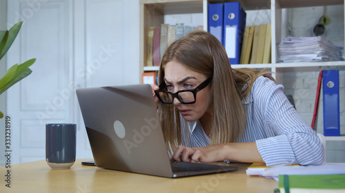 Portrait of funny goofy young woman wearing glasses trying to see what she's typing working on laptop computer in modern office. First day at work.