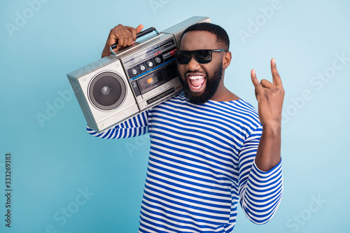 Hard rock. Photo of funny dark skin guy hold retro cassette recorder on shoulder showing metal horns hand wear sun glasses striped sailor shirt isolated blue color background