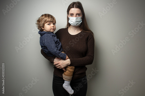 A young woman wearing a face mask and holding in her hands a toddler on a white background. Protection against the COVID-19 transmission. Mother and child in quarantine.