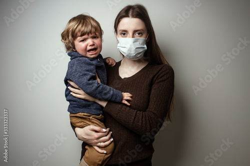 A young woman wearing a face mask and holding in her hands a crying toddler on a white background. Protective measures. Mother and child in quarantine. A young woman holding a baby boy.