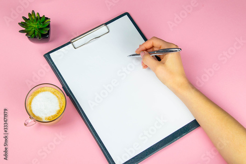 Tablet with white flattery next to which stands a green plant, hand holds a pen and writes on a sheet in the other hand a cup with a coffe